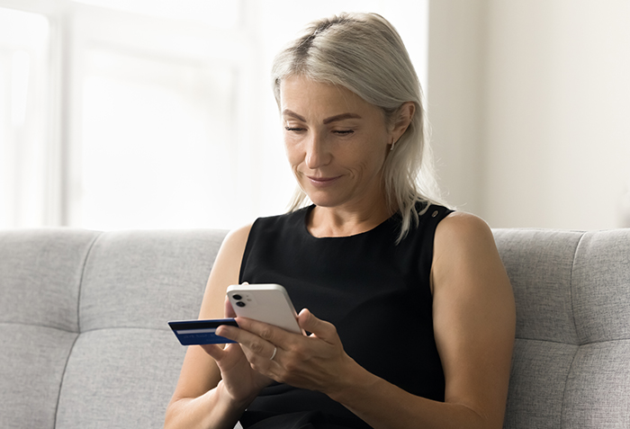 A woman sitting on a couch holding a credit card and a smartphone. She appears to be paying a bill, or checking to see whether a bill has been paid.