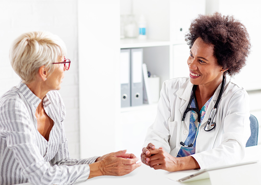 A healthcare provider in a white coat, sitting at a table with a middle-aged woman. The provider is smiling and is having a conversation with the woman.